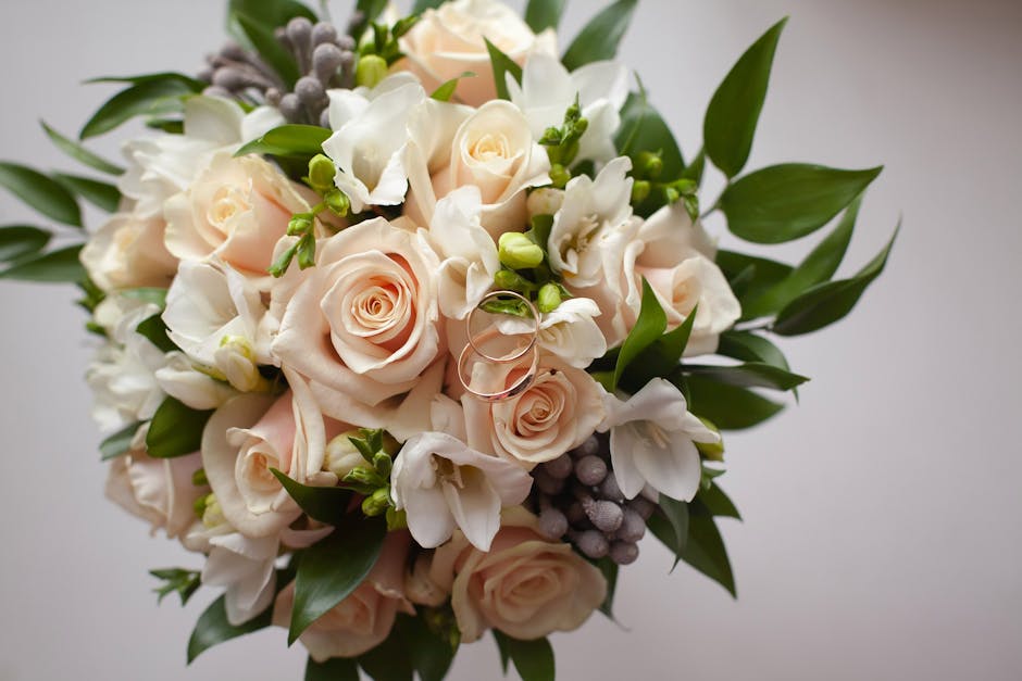A close-up view of a floral bouquet arranged with pale peach roses, white freesias, and green foliage, accentuated by small grey berries, with two wedding rings delicately placed among the flowers. Th
