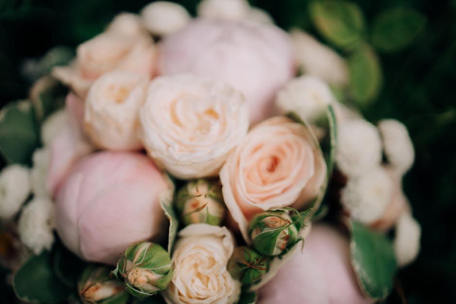 A close-up view of a bouquet of fresh, soft pink and blush-colored roses with delicate, tightly packed petals. The arrangement includes some small, round white blooms and green foliage with glossy lea