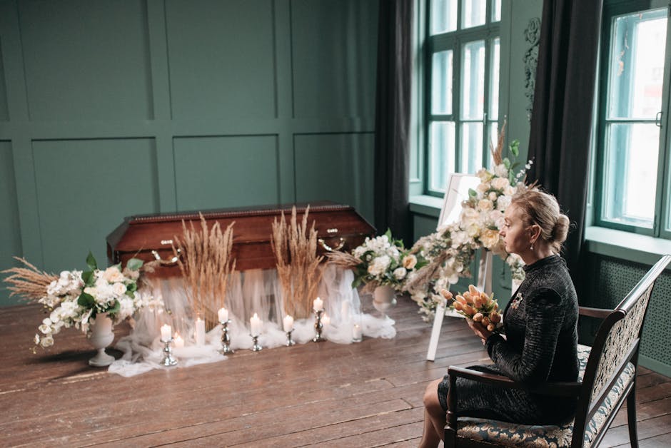 A woman seated in a dark, patterned dress holds a bouquet of peach and pink roses and greenery in her hands during a floral tribute. In front of her, on a wooden floor, is a sympathy flower arrangemen