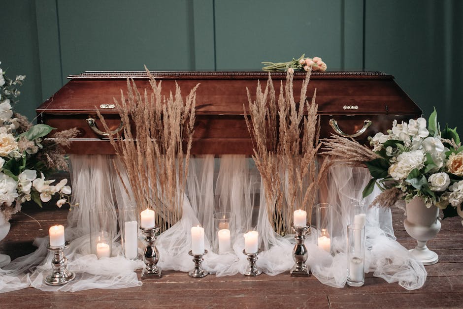 A memorial display featuring a polished wooden casket at the center, adorned with tall arrangements of dried pampas grass and soft pink roses laid on top. Surrounding the casket are multiple white can